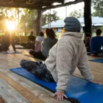 Deelnemers zitten op yogamatten tijdens een rustige buitenles onder een houten overkapping.