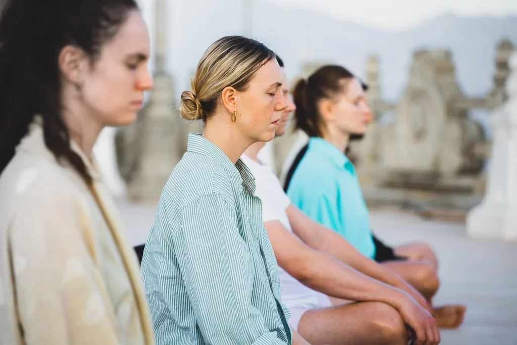 Drie mensen zitten met gesloten ogen rustig naast elkaar tijdens een meditatie in de buitenlucht.